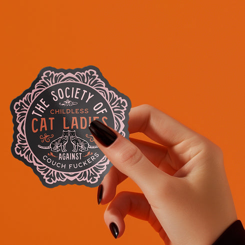 Round sticker with ornate border showing two cats and text "The Society of Childless Cat Ladies Against Couch Fuckers" in pink and purple versions on black background.